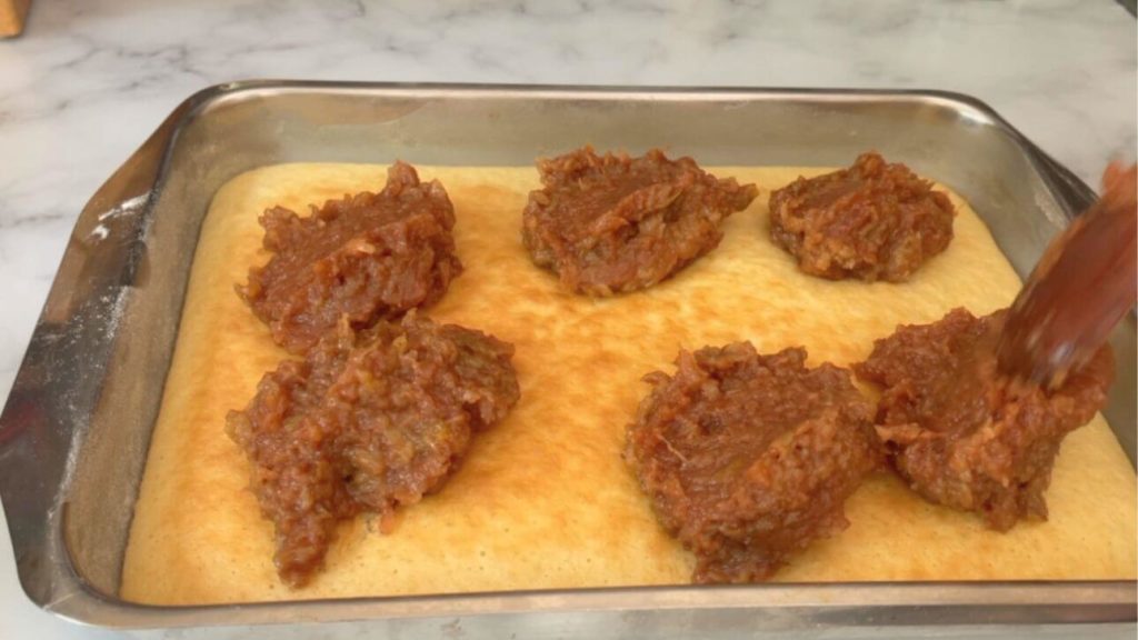 Half of the apple cake batter baked in a pan with a layer of apple filling being added on top for making a delicious apple cake.