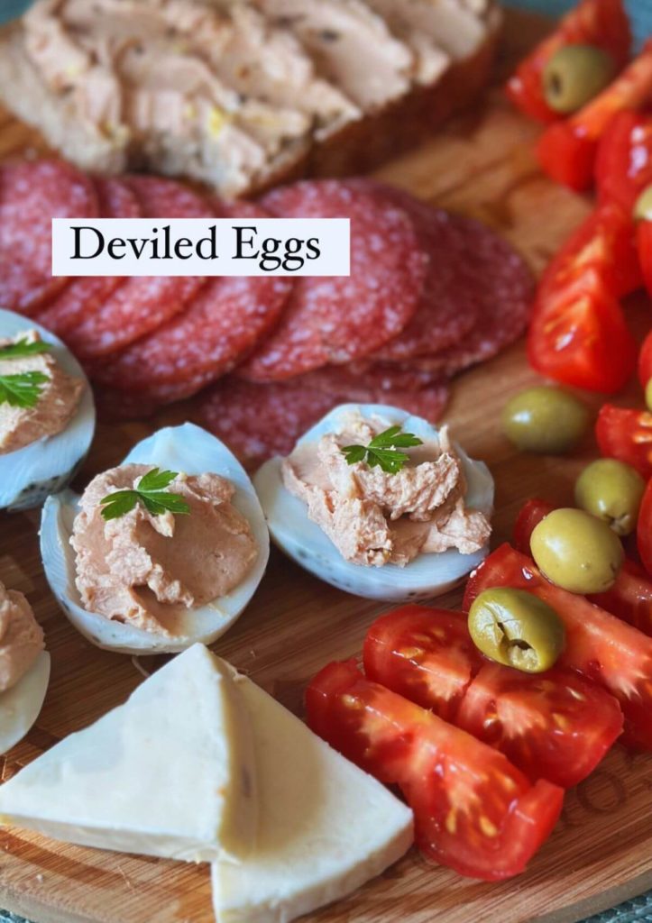 An appetizing plate with deviled eggs, salami slices, green olives, sliced tomatoes, cheese triangles, and a slice of bread with a generous egg filling spread.