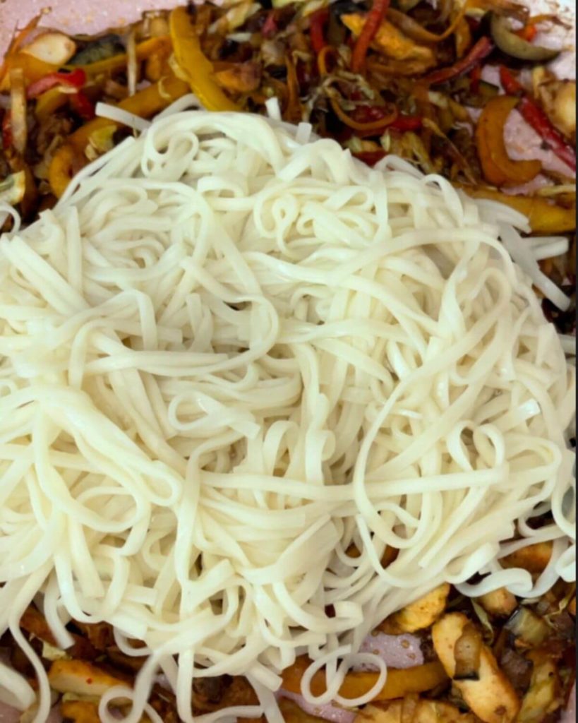 Colorful stir-fry with shredded cabbage, carrots, bell peppers, courgette, and aubergine, topped with cooked noodles in a pan.