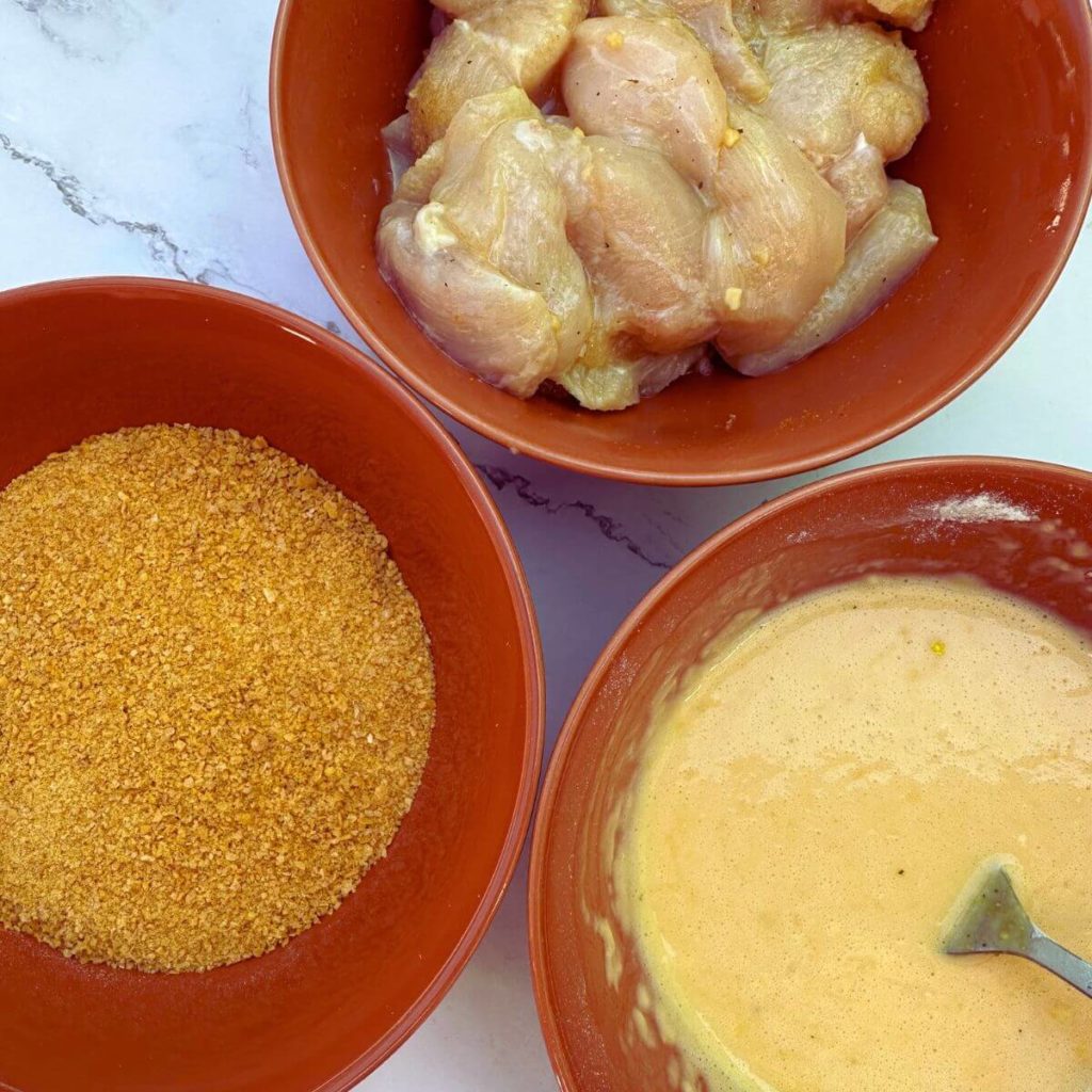 Freshly diced and seasoned chicken breast in a mixing bowl, alongside a bowl of breadcrumbs and a beaten egg mixture.