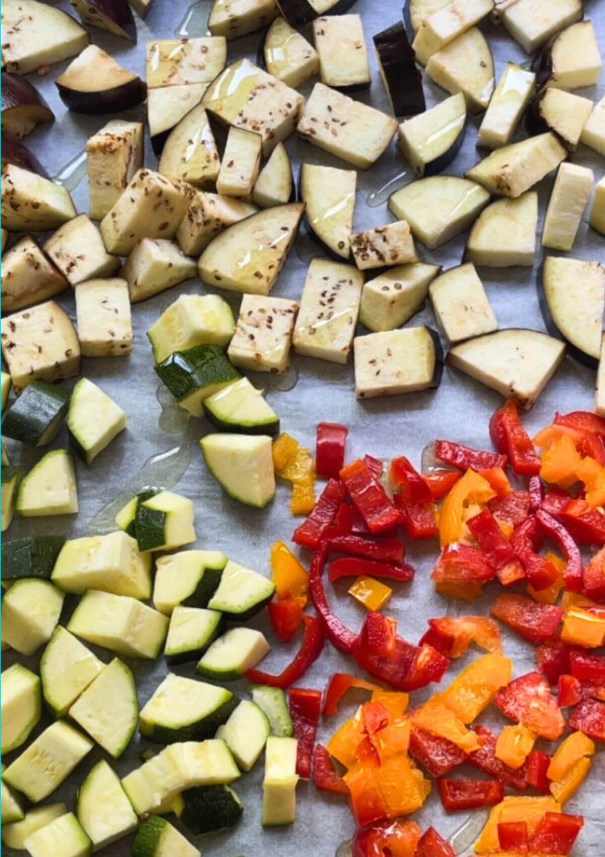 Cubed eggplant, zucchini, yellow and red bell peppers seasoned and drizzled with olive oil in a baking pan, prepared for oven grilling.