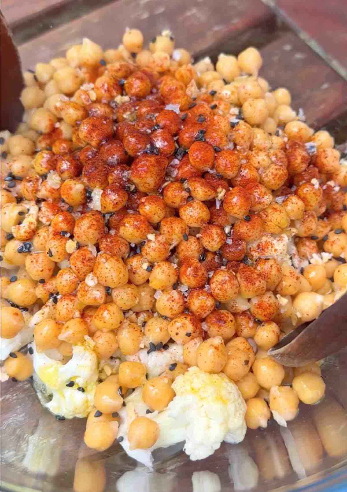 A bowl filled with cauliflower florets and chickpeas, seasoned with spices, ready to be mixed.