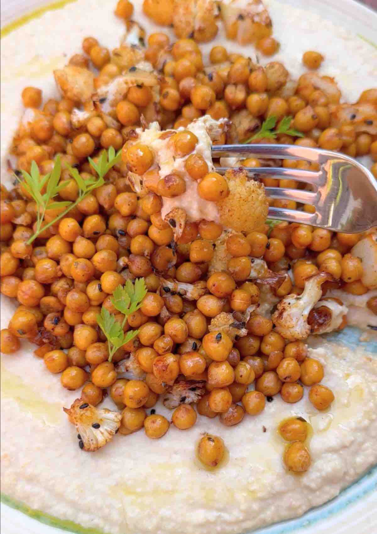 Oven roasted cauliflower and chickpeas with hummus, featuring a fork in the air with a perfect bite.