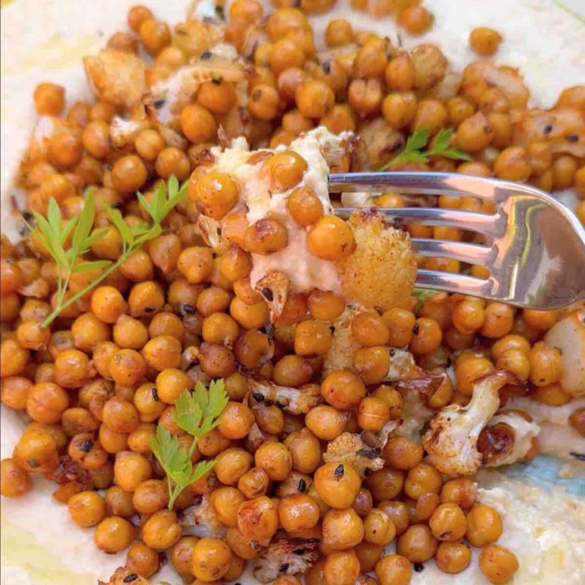 Oven roasted cauliflower and chickpeas with hummus, featuring a fork in the air with a perfect bite.