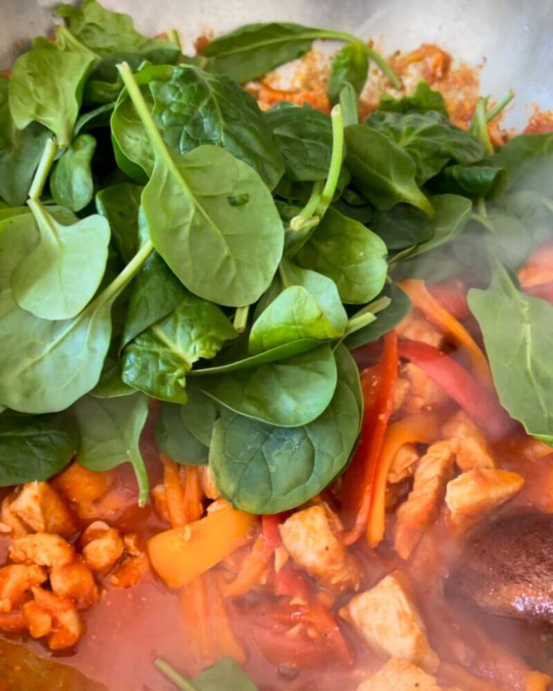Chicken breast pieces cooked in a pan with colorful peppers cut lengthwise, tomato sauce and fresh spinach.