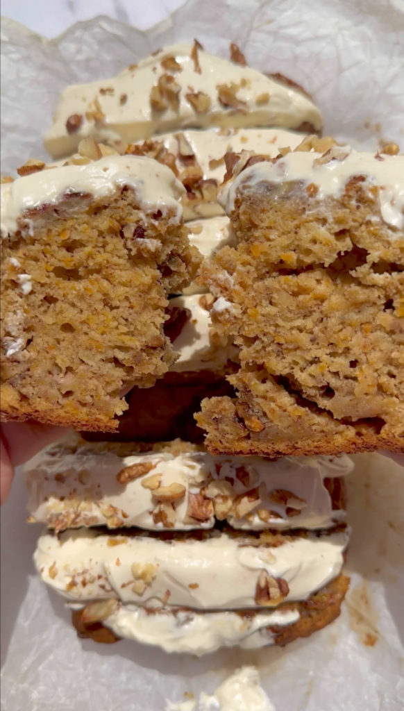 Vegan carrot cake banana bread topped with cream cheese frosting and walnuts, held in hand with a slice torn in half.