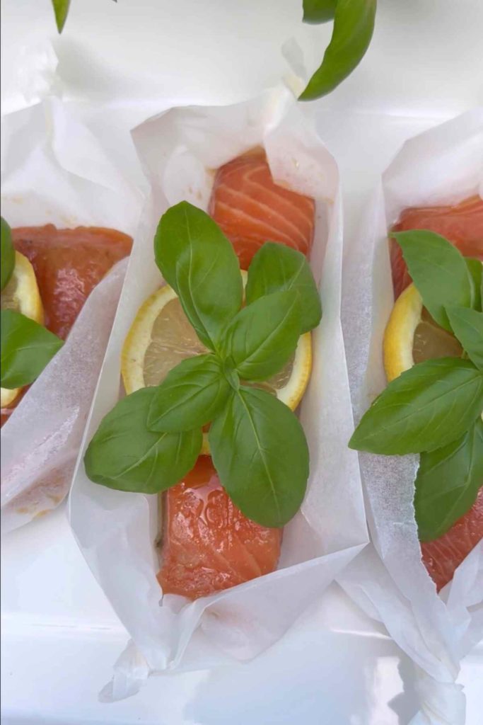 Salmon fillets marinated with lemon slices and adorned with fresh basil leaves, neatly nestled in parchment paper for oven baking.