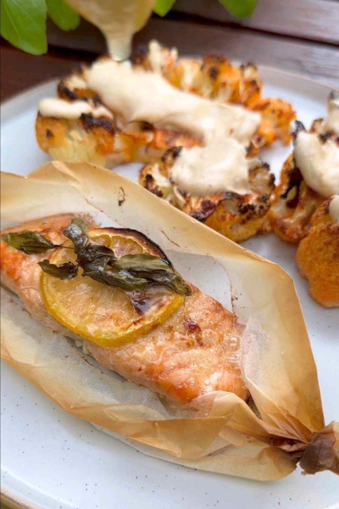 Salmon fillets marinated and baked with lemon slices and fresh basil atop, enveloped in parchment paper, accompanied by cauliflower steaks.