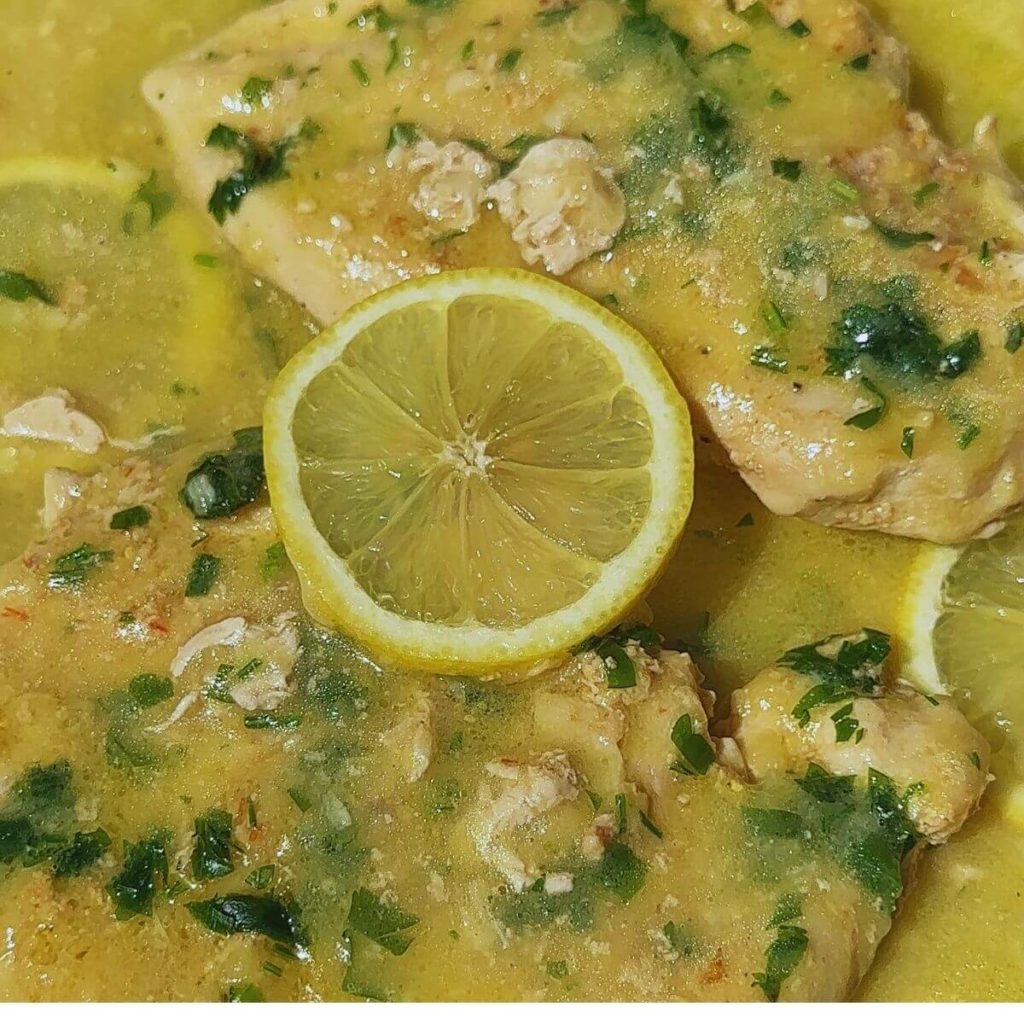 Two succulent lemon-infused chicken breast fillets with a zesty lemon sauce, garnished with fresh lemon slices and vibrant green herbs, beautifully presented in a pan.