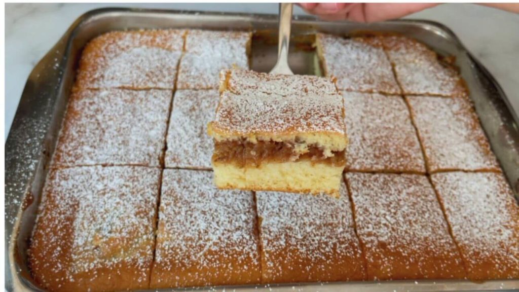 A slice of apple cake being held over a rectangular pan filled with apple cake squares, delicately dusted with powdered sugar for a sweet finishing touch.