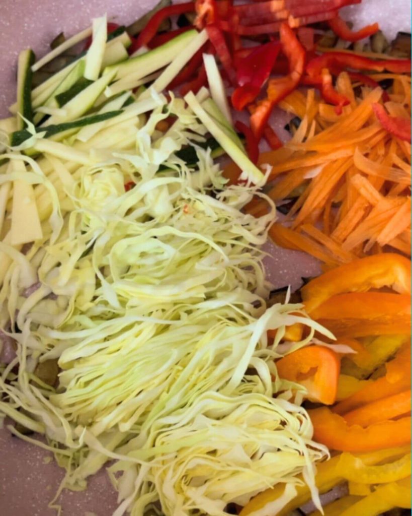 Pan filled with a colorful mix of shredded cabbage, carrot, red and yellow bell pepper, green bell pepper, courgette, and aubergine cut lengthwise
