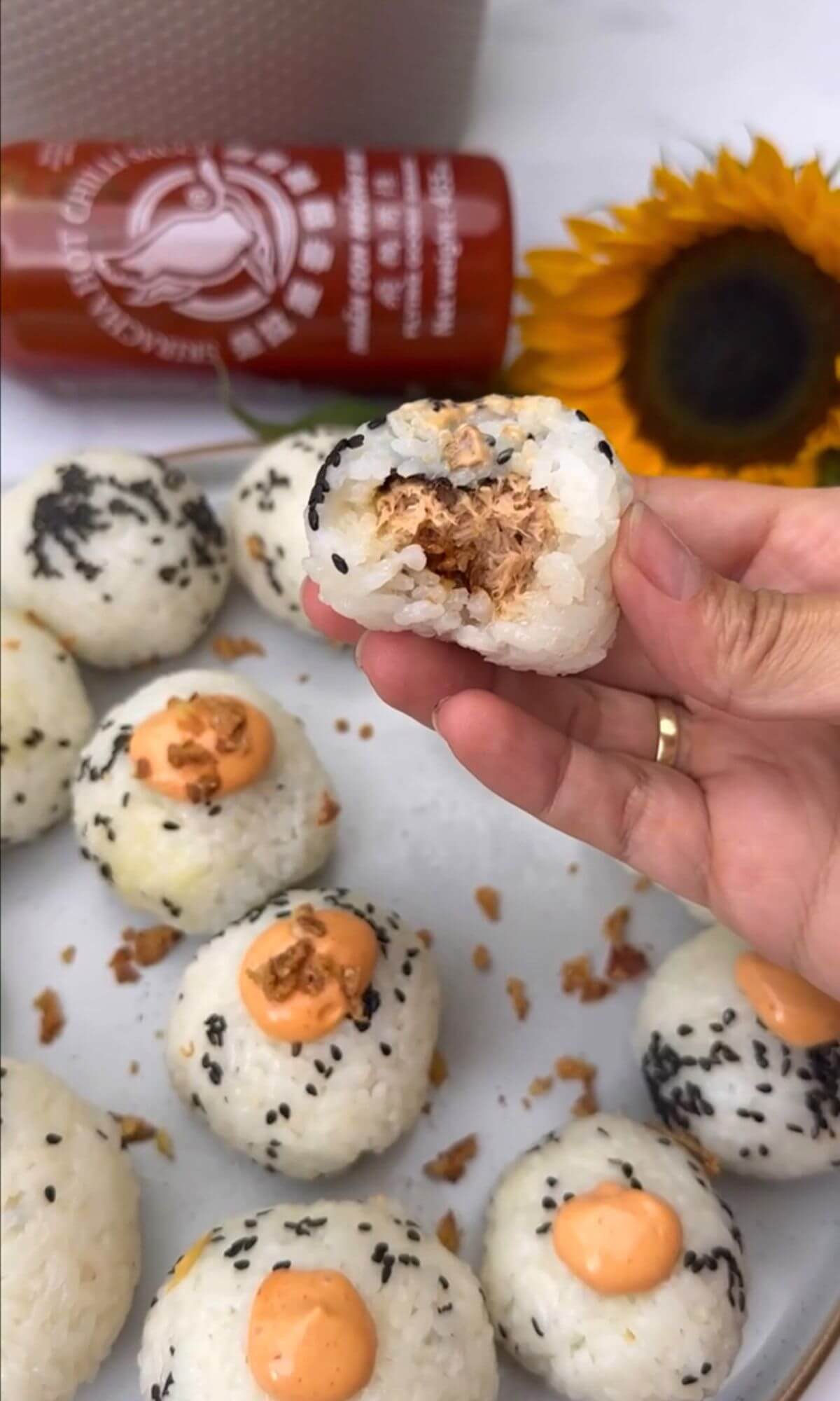 The image depicts a hand holding a rice ball filled with tuna in the center, with sesame seeds sprinkled on top, and bitten in half. In the background, there is a plate of tuna onigiri, a bottle of sriracha sauce, and a sunflower.