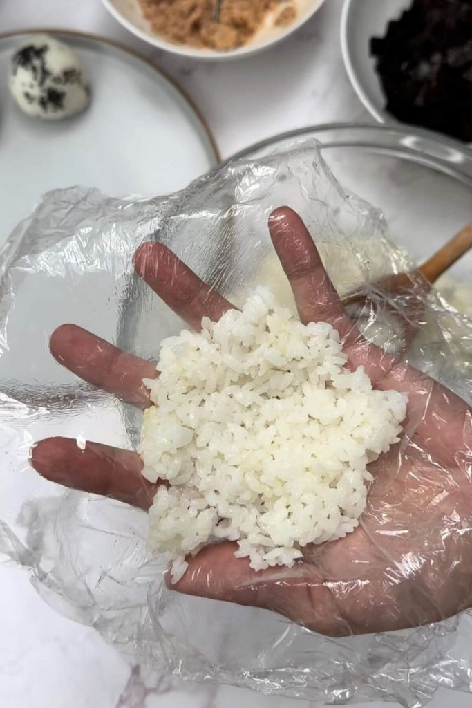 In the picture, there's a table with different bowls. One has tuna, another has rice, and there's also a bowl with seaweed cut into small pieces. On a plate, there's a rice ball called onigiri with tuna inside. In the middle, a hand is holding a square piece of plastic wrap with cooked rice on it.
