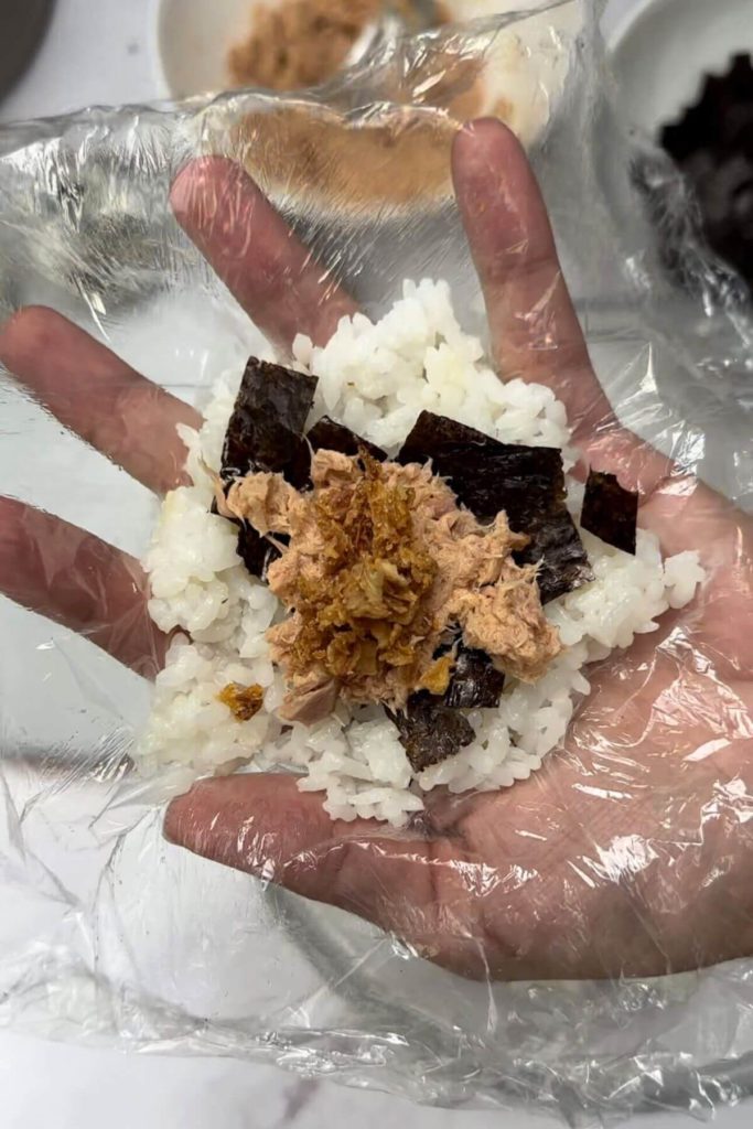 In the picture, there's a table with bowls of tuna, rice, and seaweed cut into small pieces. There's also a plate with a special Japanese rice ball called onigiri with tuna inside. In the middle, someone's holding a square piece of plastic wrap with cooked rice, seaweed pieces, and a spoonful of tuna mixed with onion flakes on top.