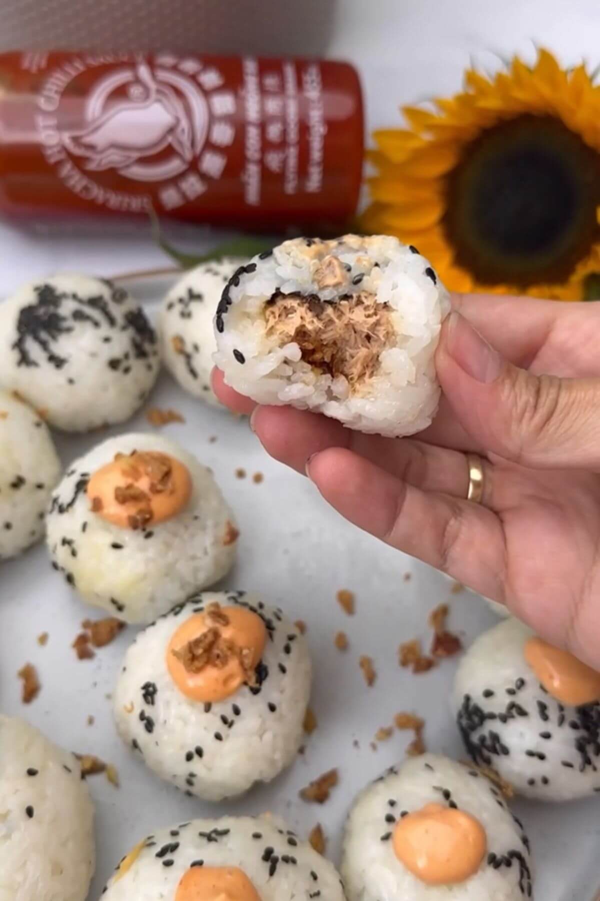 The image depicts a hand holding a rice ball filled with tuna in the center, with sesame seeds sprinkled on top, and bitten in half. In the background, there is a plate of tuna onigiri, a bottle of sriracha sauce, and a sunflower.