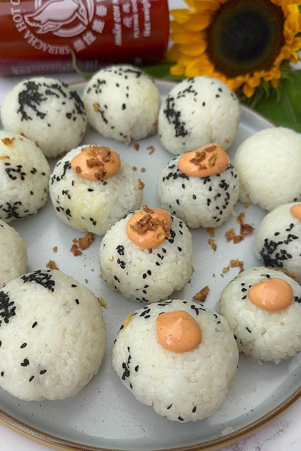 The picture shows a plate with tuna rice balls topped with sesame seeds, a little bit of spicy mayo, and onion flakes. There's also a bottle of sriracha sauce and a sunflower next to it.