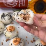 Hand holding spicy tuna onigiri, sesame crust, with creamy filling visible.