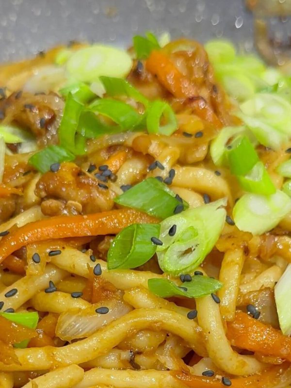 Udon noodles with teriyaki chicken in a pan, topped with green onions and sesame seeds.