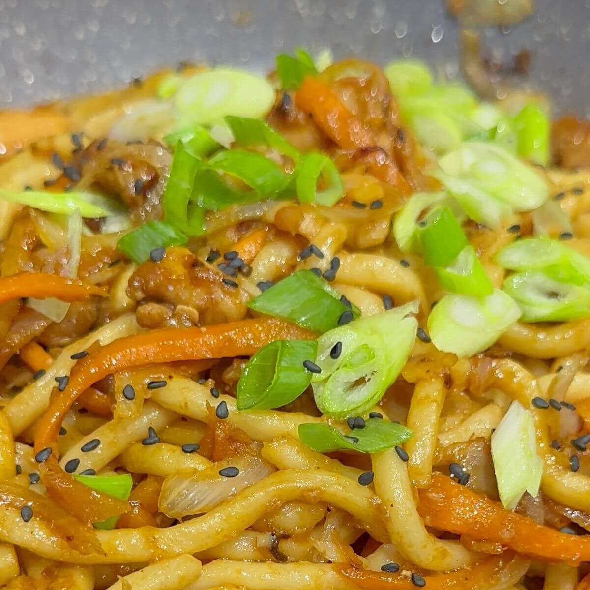 Udon noodles with teriyaki chicken in a pan, topped with green onions and sesame seeds.