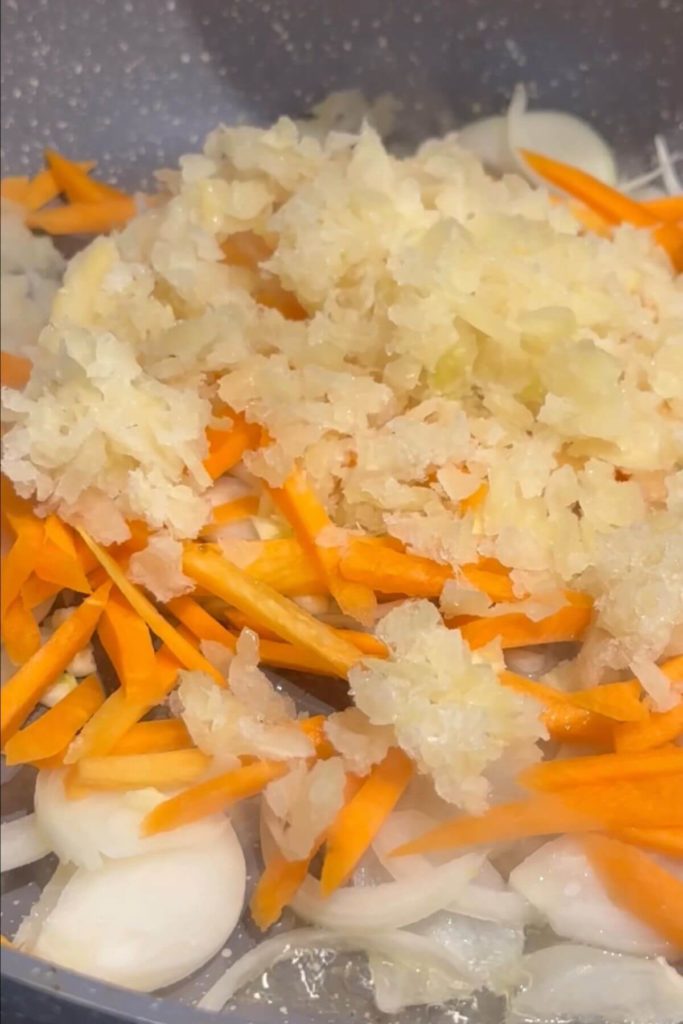 Fresh vegetables sizzling in a pan: sliced white onion, carrots, diced garlic, and shredded white cabbage being stir-fried. 