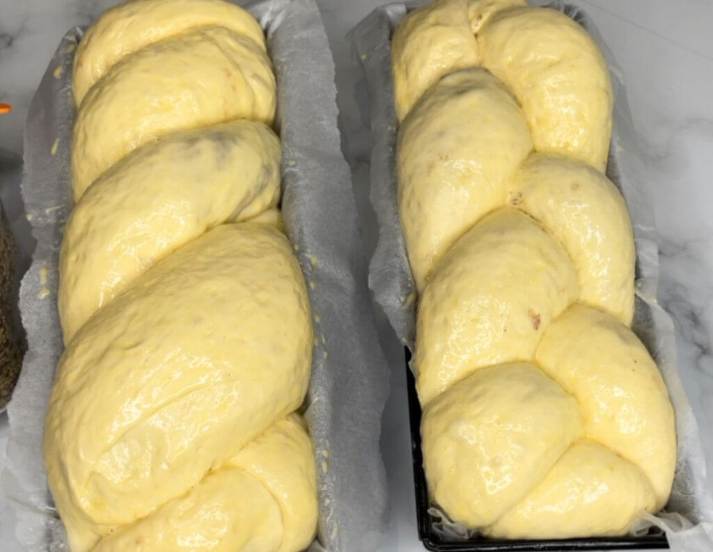 Two rolls sitting in pans lined with paper. One roll is made with 3 braids and the other with 2 braids. They're all puffed up and ready to bake in the oven.