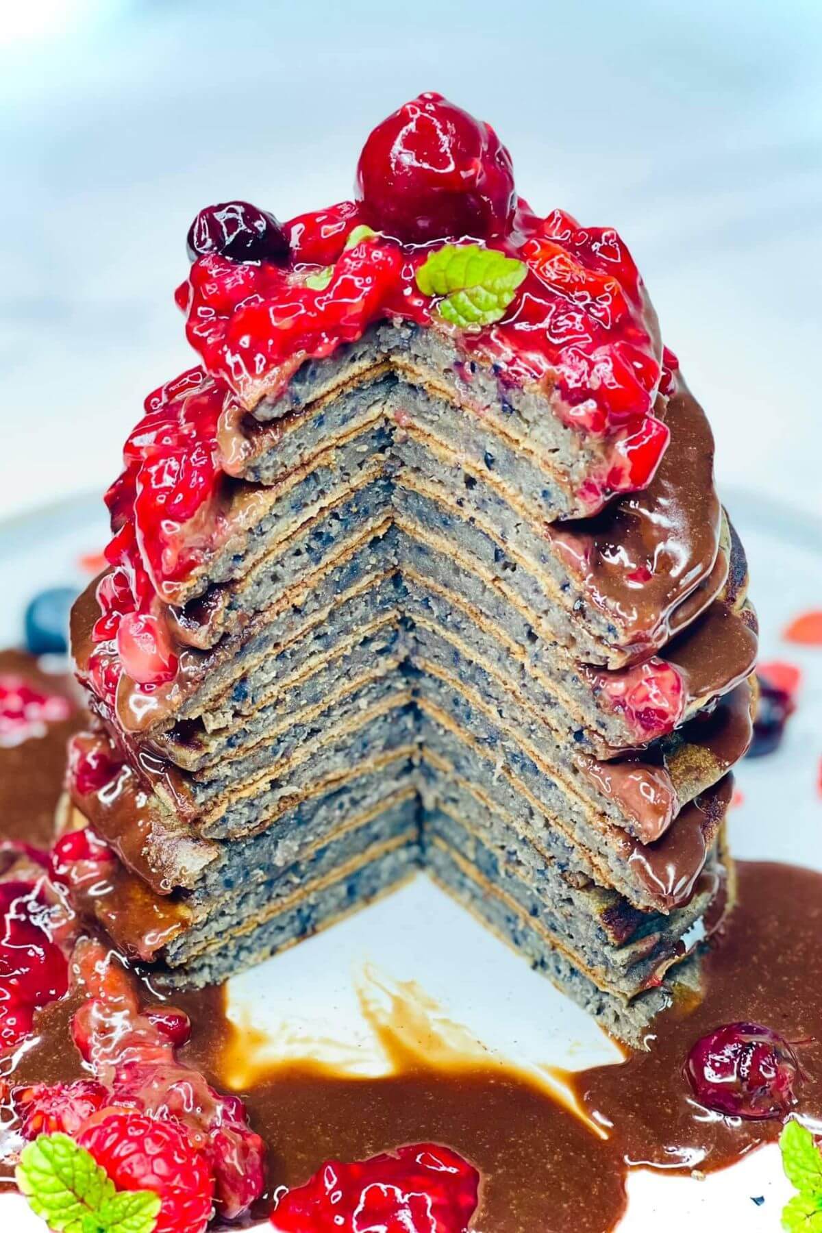 Stack of berry pancakes on a plate with mixed berry sauce and chocolate ganache drizzle, with a wedge cut out to show the interior fluffy texture. 