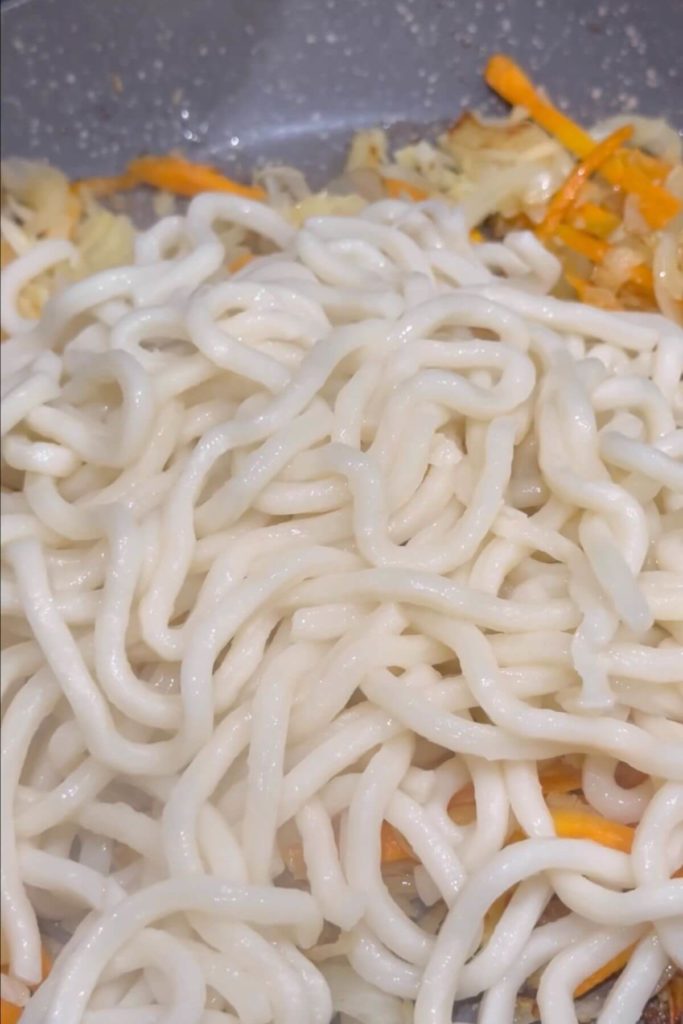 Stir-frying sliced white onion, carrots, diced garlic, and shredded white cabbage in a pan, then adding cooked udon noodles.