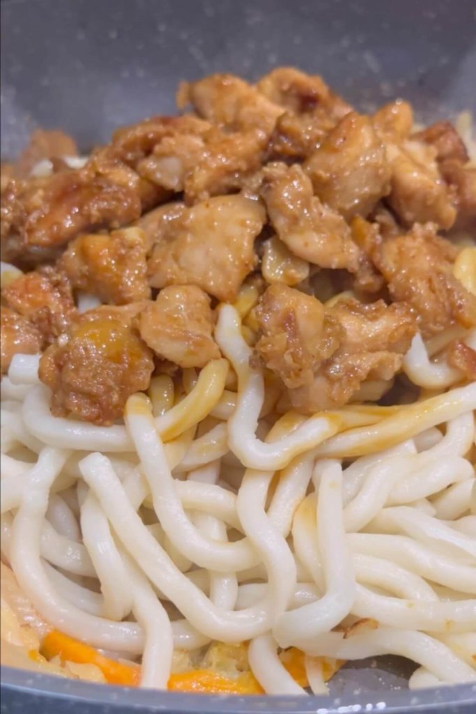 Stir-fried sliced white onion, carrots, diced garlic, and shredded white cabbage in a pan, followed by cooked udon noodles and a sauce made of dark soy sauce, oyster sauce, mirin, brown sugar, and red pepper flakes. Finished with cooked chicken teriyaki.