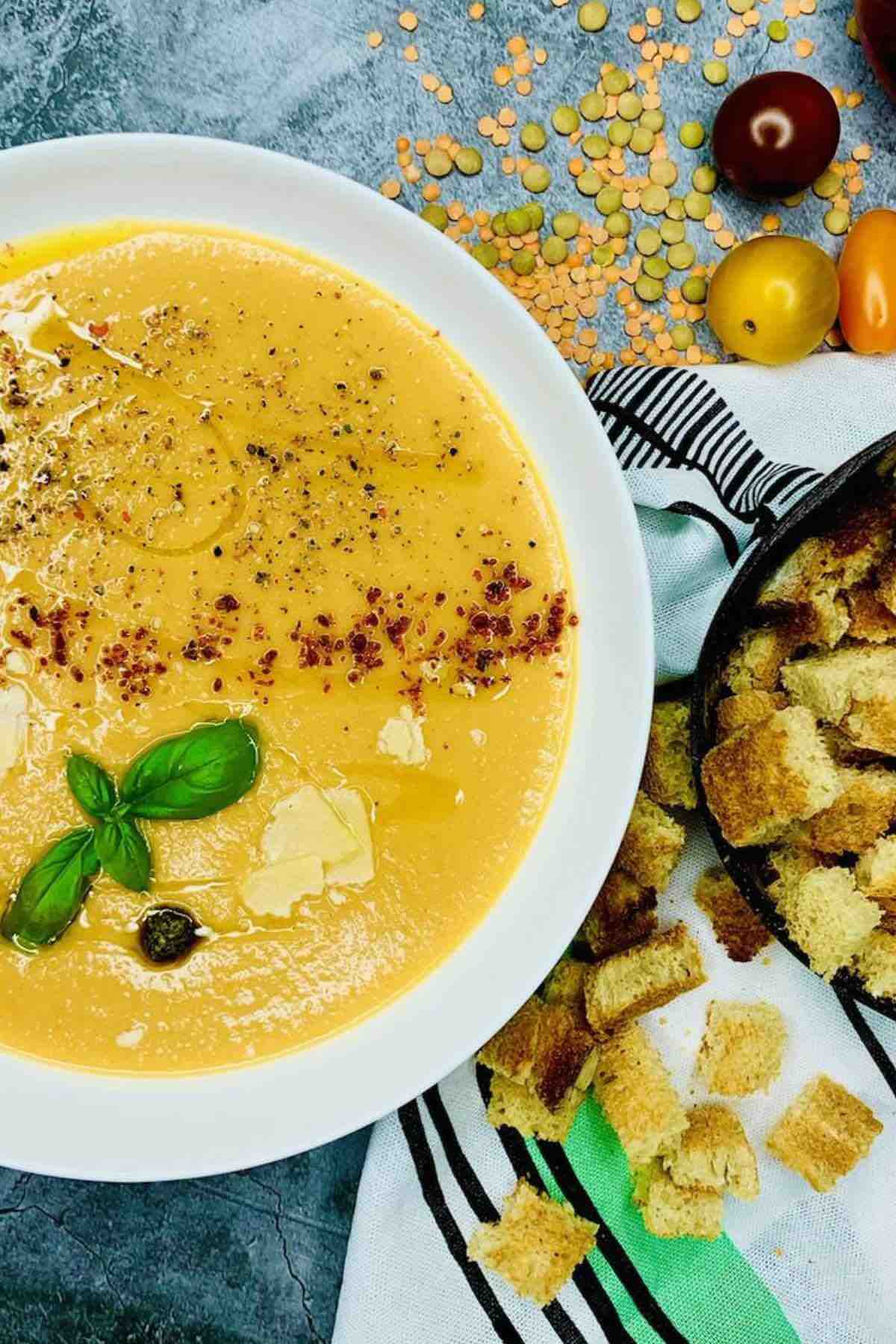Creamy vegan lentil soup with chili powder, parmesan, basil, croutons, and dry yellow lentils in the background.