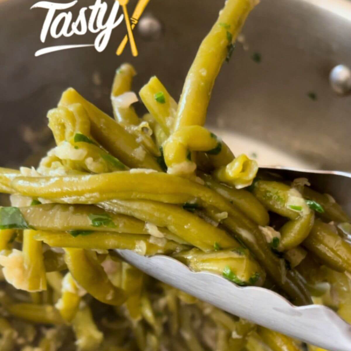 Sautéed green beans with garlic, garnished with chopped parsley, being lifted with kitchen tongs.