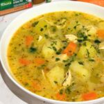 A bowl of chicken soup with herbs, surrounded by fresh vegetables.