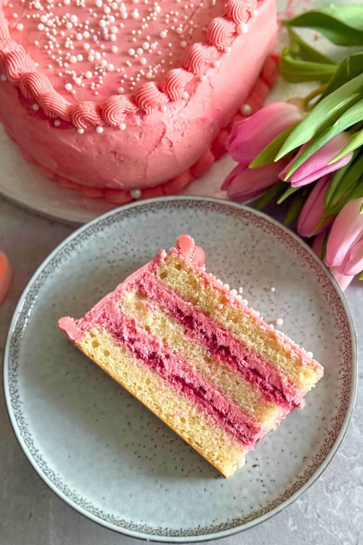 A slice of Raspberry Heart Shaped Cake on a grey plate with some pink tulips in the background and a part of the whole cake.
