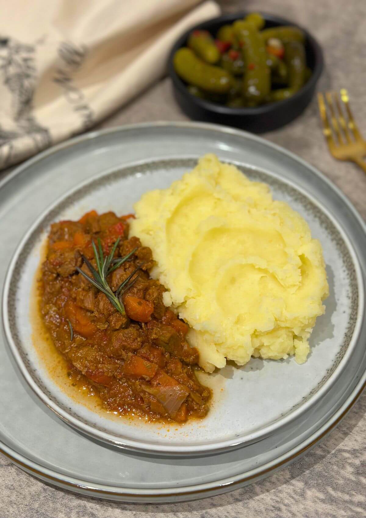 Hearty Beef Stew with Creamy Mashed Potatoes on a grey plate with pickled cucumbers in the background.
