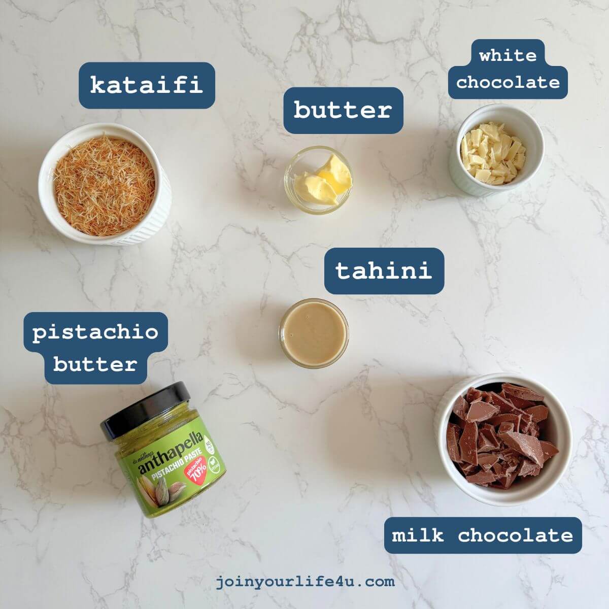 White and milk chocolate, butter, kataifi (dried and chopped), pistachio butter, tahini individually displayed on a table. 