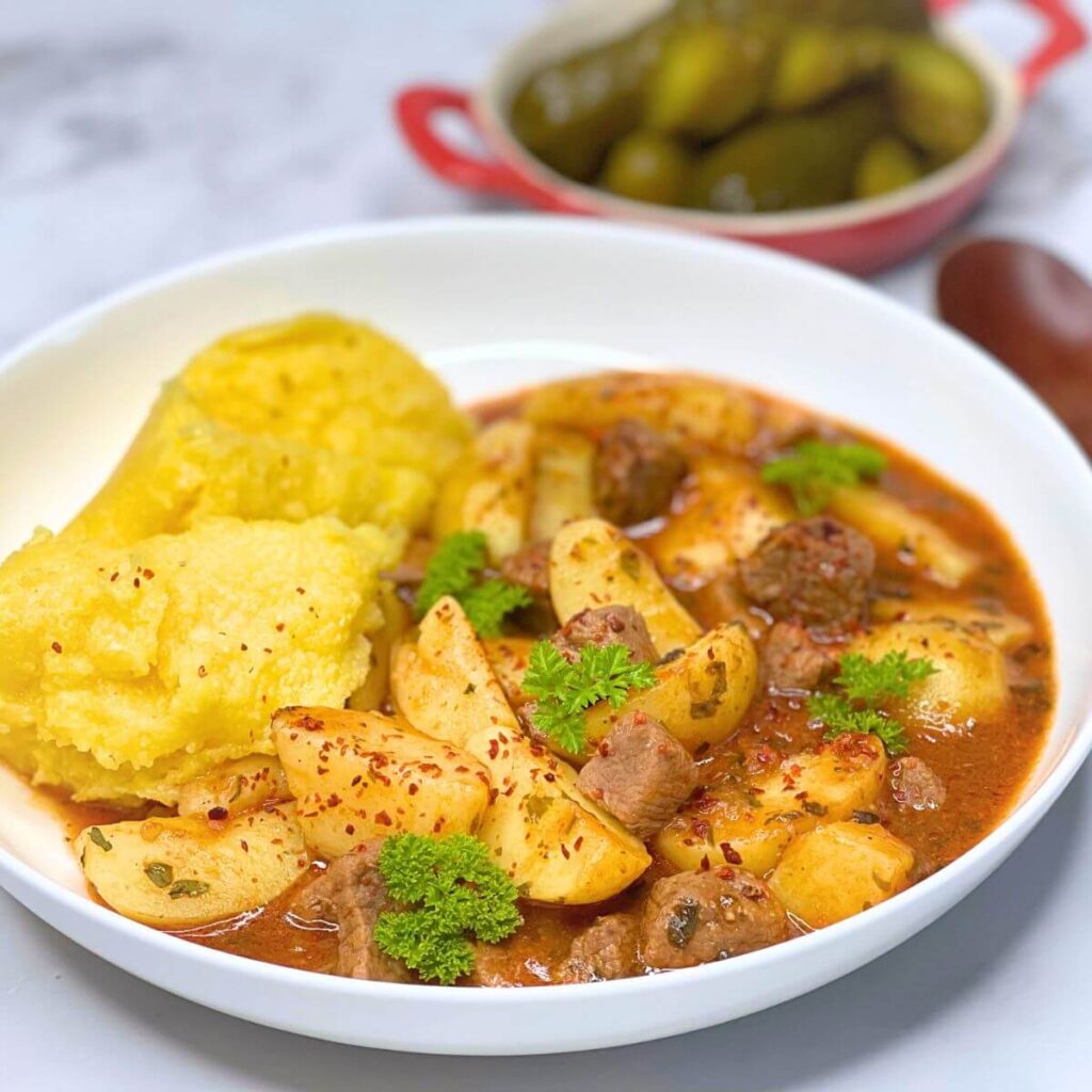 Bowl of beef potato stew with a spoon of polenta on the side, garnished with curly parsley. 