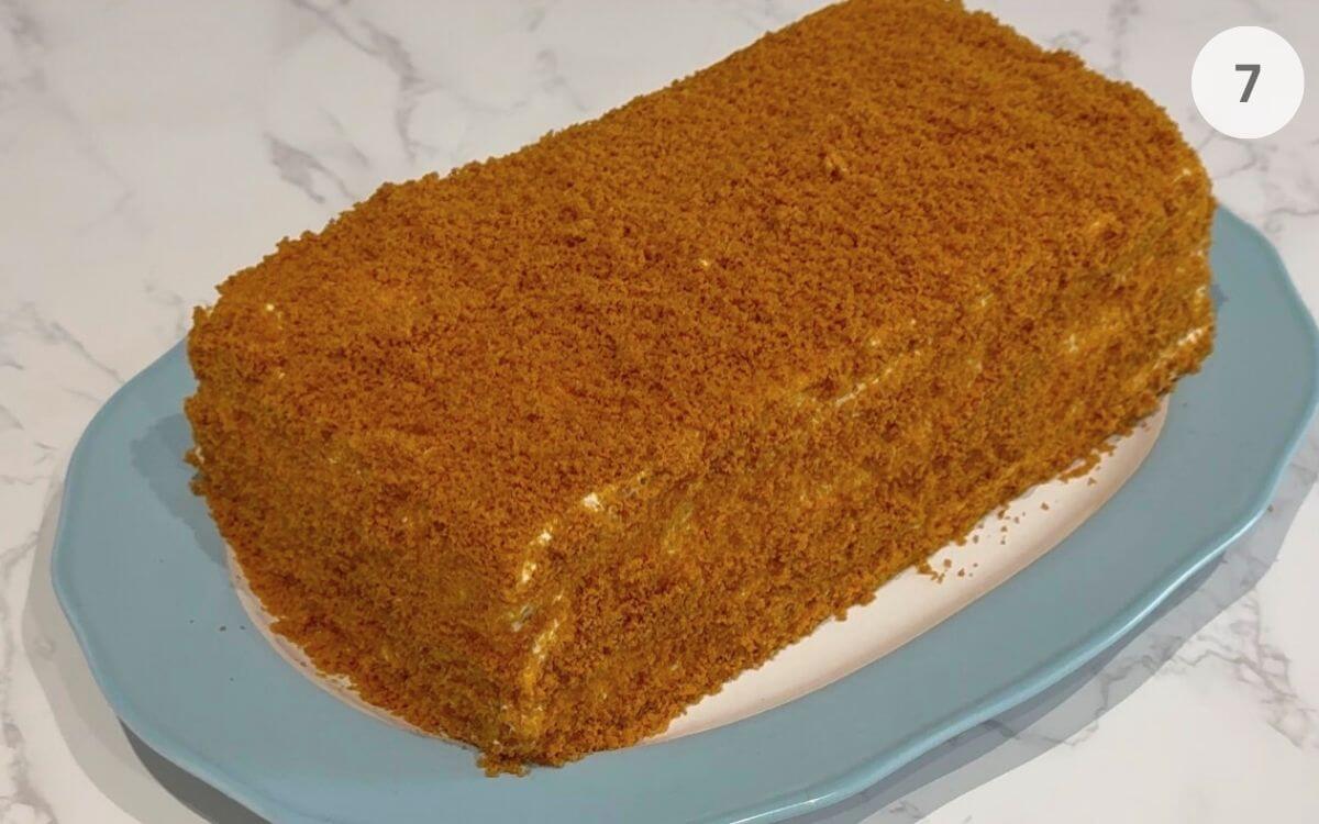 A traditional Russian Medovik cake fully coated in golden honey cake crumbs on a serving plate.