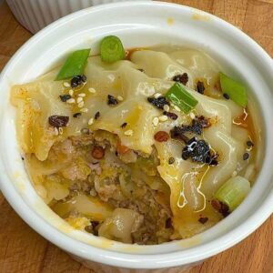 Steamed pork dumpling lasagna in white ramekin topped with green onions sesame seeds and chili oil.