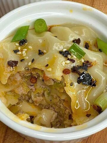 Steamed pork dumpling lasagna in white ramekin topped with green onions sesame seeds and chili oil.