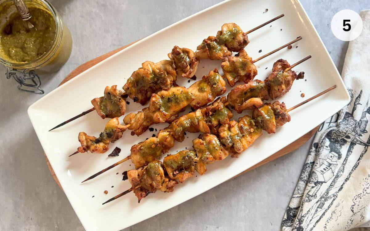 Grilled chicken satay skewers drizzled with bright green Thai basil pesto on white platter.