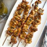 Grilled chicken satay skewers drizzled with green Thai basil pesto sauce on a white platter.
