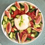 A white serving bowl containing sliced purple figs, crisp green cucumbers, ripe tomatoes, and creamy burrata cheese.