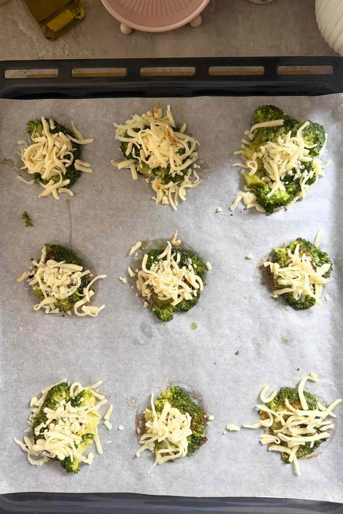 Raw smashed broccoli florets flattened on a baking sheet, topped with shredded parmesan and sprinkled with spices.