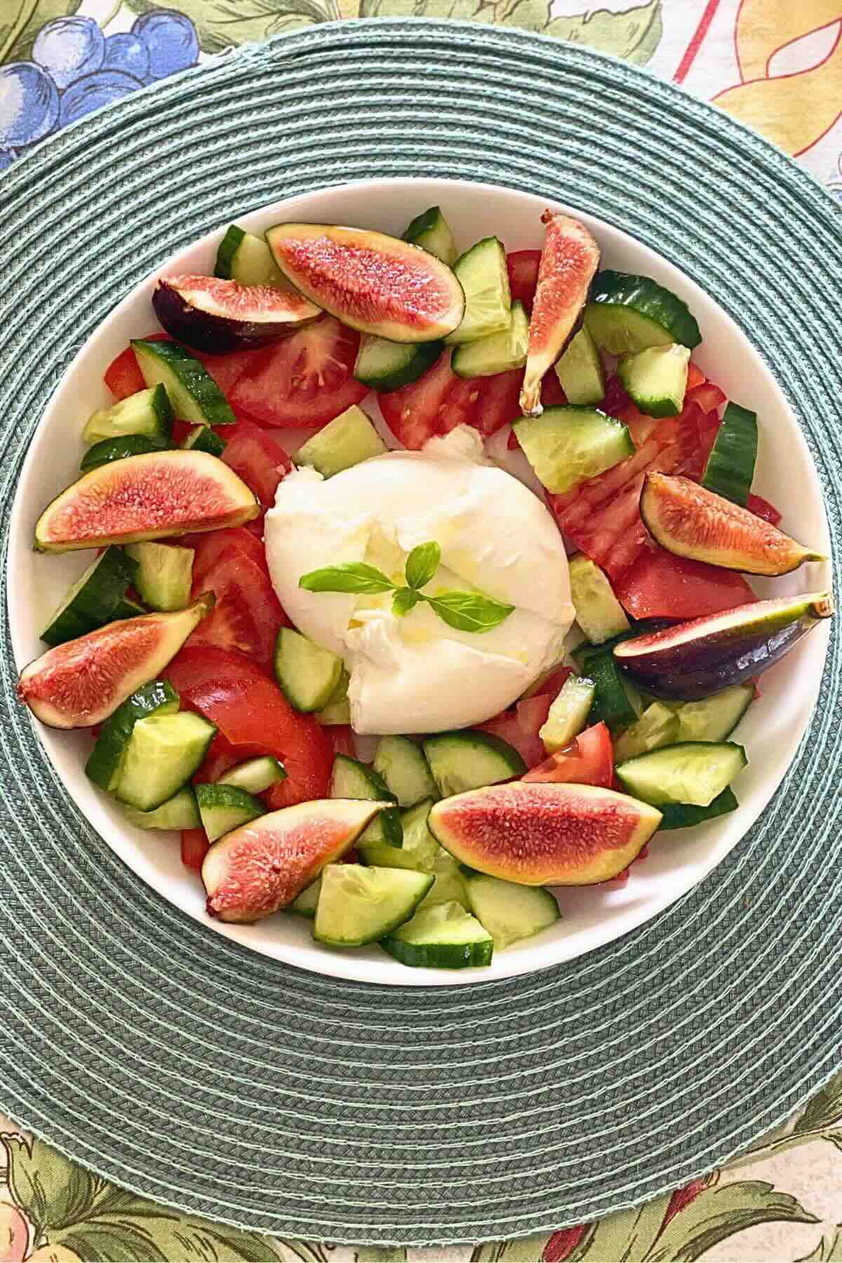 A white serving bowl containing sliced purple figs, crisp green cucumbers, ripe tomatoes, and creamy burrata cheese.