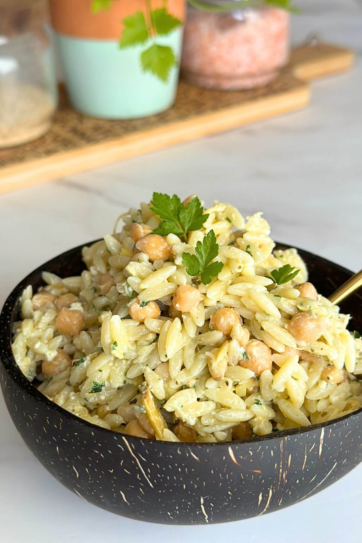 Bowl of cold orzo salad with chickpeas, pine nuts, artichoke hearts, and feta cheese on a wooden table.