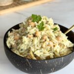 Bowl of cold orzo salad with chickpeas, pine nuts, artichoke hearts, and feta cheese on a wooden table.