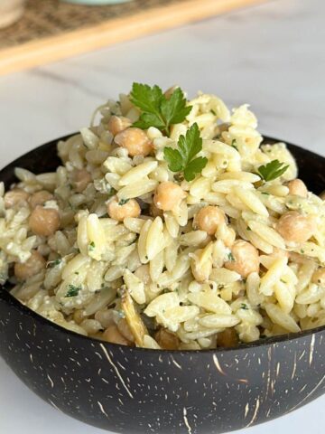 Bowl of cold orzo salad with chickpeas, pine nuts, artichoke hearts, and feta cheese on a wooden table.
