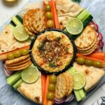 Fresh raw vegetables olives and biscuits around a bowl of chickpea dip on platter.