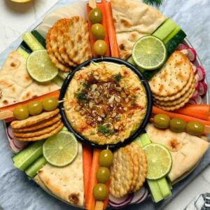 Fresh raw vegetables olives and biscuits around a bowl of chickpea dip on platter.