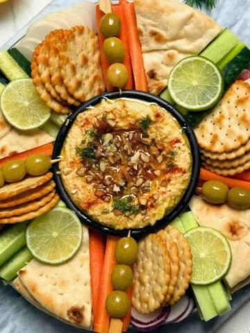 Fresh raw vegetables olives and biscuits around a bowl of chickpea dip on platter.