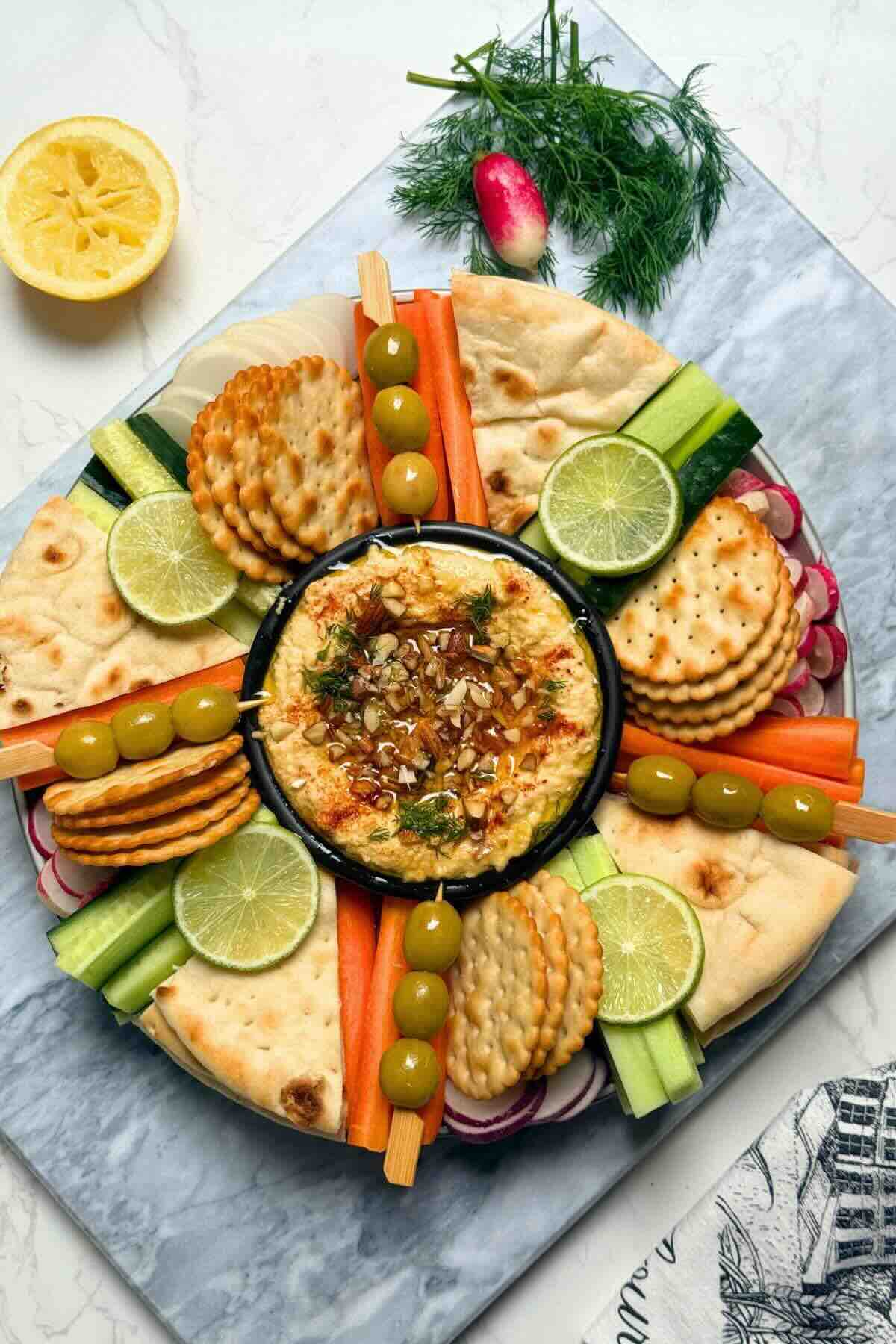 Fresh raw vegetables olives and biscuits around a bowl of chickpea dip on platter.